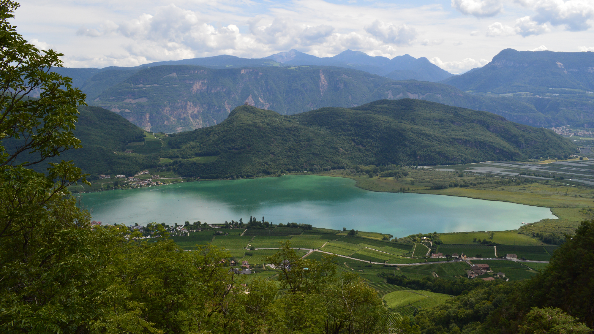 Panoramafoto: Der Kalterer See von einer erhöhten Position aufgenommen. Vor dem See Felder, einige Gebäude sowie eine Straße; hinter dem See weitere Felder sowie bewaldete Berge